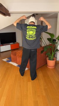 Czarna koszulka z żywym zielonym wzorem graficznym i napisem, w połączeniu z luźnymi czarnymi spodniami. Biała czapka beanie i wnętrze z rośliną.