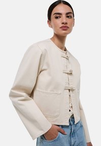Veste cropped de couleur crème avec un col rond, quatre boutons à bascule sur le devant et deux poches avant. Le tissu a une finition texturée.