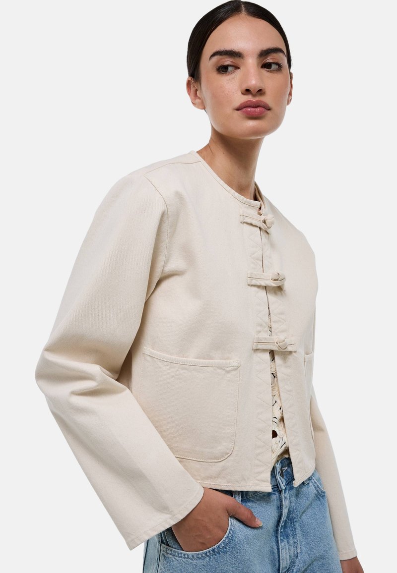 Veste cropped de couleur crème avec un col rond, quatre boutons à bascule sur le devant et deux poches avant. Le tissu a une finition texturée.