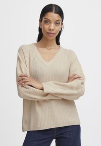 Beige V-nyakú pulóver bordázott textúrával, hosszú ujjakkal és laza fazonnal, sötétkék farmerral párosítva. Puha anyagból készült.