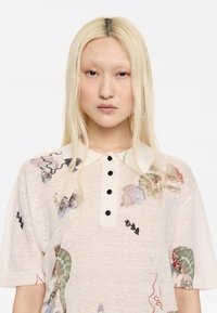 Polo shirt a maglia color crema con maniche corte, caratterizzato da un colletto con bottoni, motivi floreali astratti in vari colori e una consistenza morbida.