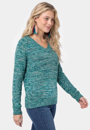 Una mujer rubia luce un jersey turquesa estampado con escote en V, unos vaqueros azules y unos pendientes largos y multicolores sobre un fondo liso y claro.