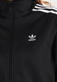 Chaqueta negra con cremallera hecha de tela texturizada, que presenta tres rayas blancas en los hombros y un pequeño logotipo blanco de Adidas en el pecho.