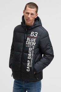 Man met kort haar die een zwarte kapjas draagt met witte en rode tekst, handen in de zakken, kijkend naar de zijkant tegen een effen achtergrond.