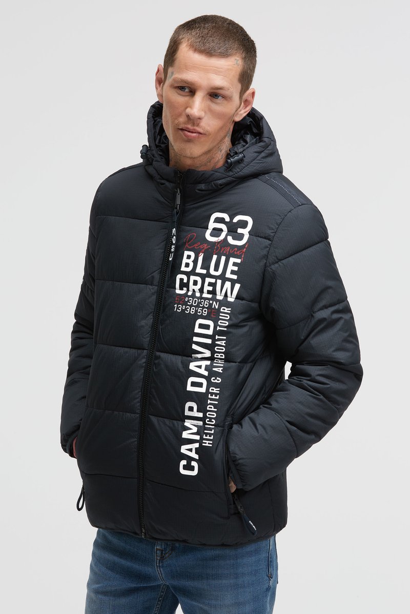 Man met kort haar die een zwarte kapjas draagt met witte en rode tekst, handen in de zakken, kijkend naar de zijkant tegen een effen achtergrond.