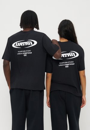 Deux personnes dos à la caméra portant des T-shirts noirs avec un texte et un logo blancs "Yourturn Studio Urban Streetwear".