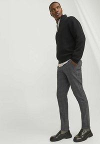 Schwarze Zip-Up-Strickjacke, graue Karohose, schwarze Slipper. Die Materialien scheinen aus Strick- und Webstoff zu sein. Lässiges, aber maßgeschneidertes Design.