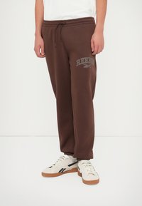 Pantalones de chándal marrones con una cinturilla ajustable, con un logo gris de Reebok en el muslo izquierdo y puños acanalados en los tobillos.