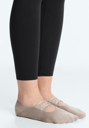 Nohy v béžových baletných ponožkách s elastickými krížovými pásmi, v kombinácii s čiernymi legínami končiacimi nad členkami.