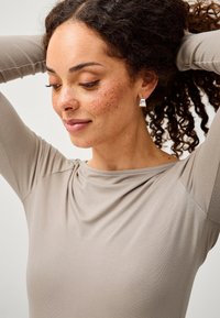 Silberne geometrische Steckerohrringe mit glatter Oberfläche, getragen von einer Person mit lockigem Haar. Beigefarbene Langarmoberteil mit drapiertem Ausschnitt und weicher Textur.