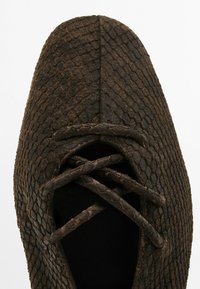Scarpa marrone con texture realizzata in materiale simile a rettile, caratterizzata da una punta affusolata e una chiusura lacci incrociati. Finitura opaca senza dettagli visibili.