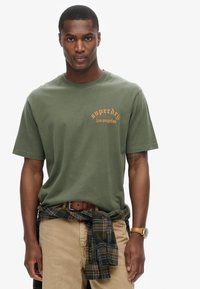 Camiseta de algodón verde oliva con logo naranja de "superdry los ángeles", de manga corta y cuello redondo, combinada con pantalones caqui y una camisa de cuadros atada a la cintura.