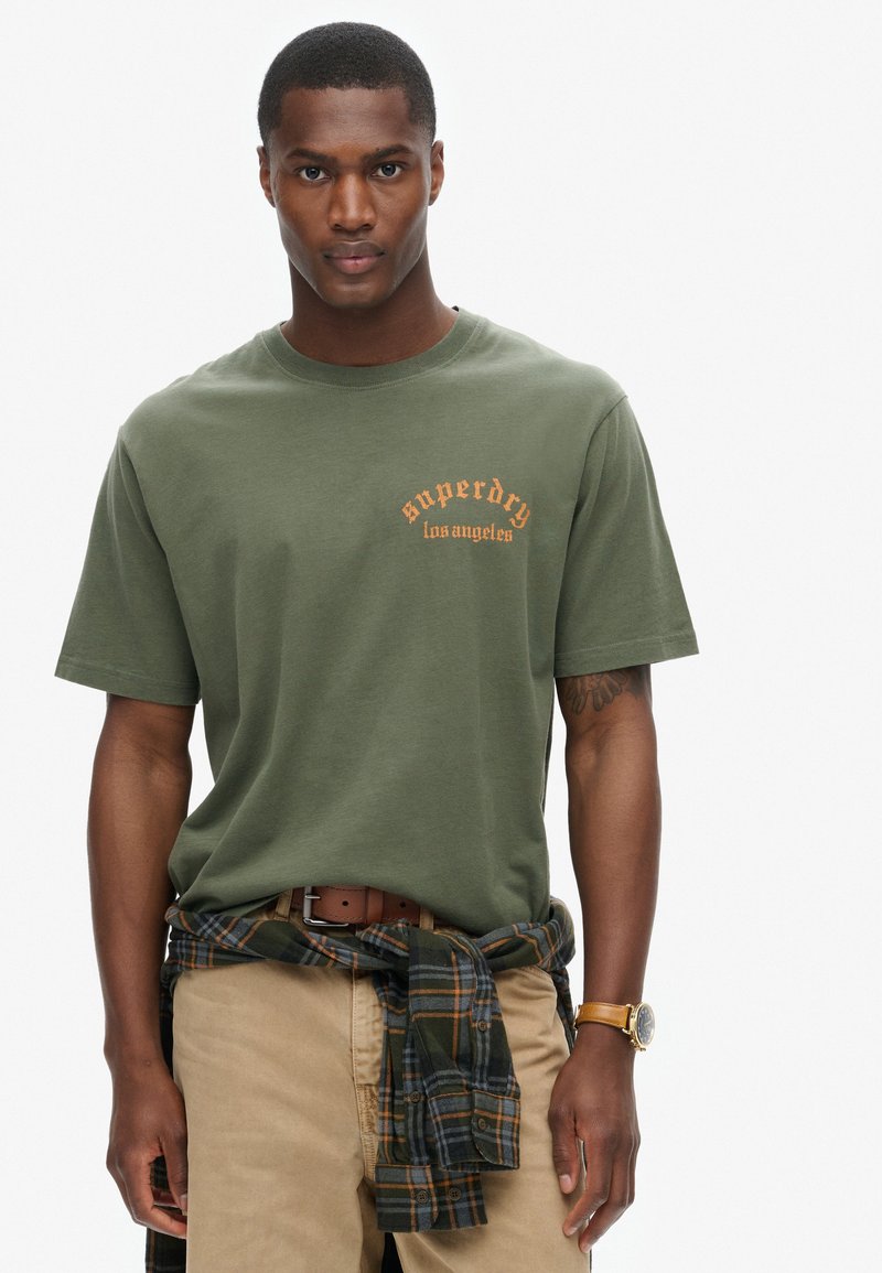 Camiseta de algodón verde oliva con logo naranja de "superdry los ángeles", de manga corta y cuello redondo, combinada con pantalones caqui y una camisa de cuadros atada a la cintura.