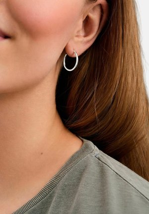 Prim-plan al urechii unei femei cu o cercel mic argintiu, păr șaten deschis și guler de cămașă gri cu dungi văzut parțial.