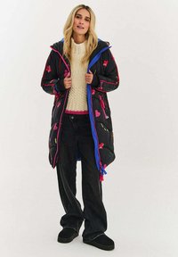 Černý péřový kabát s barevnými vyšívanými vzory, modrým zipem a kapucí. Kombinován s krémovým pleteným svetr a černé kalhoty.