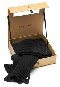Black ribbed knit beanie and scarf set inside an open cardboard gift box with "Be gentle..." printed on the lid interior.