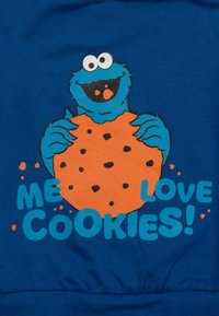 Personaggio blu con grandi occhi che tiene un grande biscotto al cioccolato arancione, con il testo "IO ADORO I BISCOTTI!" su un tessuto blu.