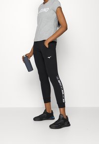 Camiseta gris con el logo "Mizuno", pantalones joggers negros con rayas blancas en los lados y zapatillas deportivas negras. Sosteniendo una botella de agua gris.