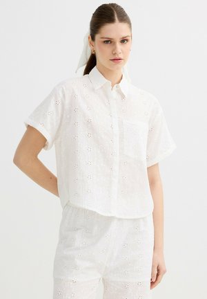 Mujer joven con el pelo recogido, vestida con una camisa blanca bordada de manga corta abotonada y pantalones cortos a juego sobre un fondo liso.