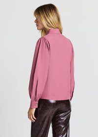 Blusa rosa con colletto alto, maniche a sbuffo e polsini con bottoni. Indossata con pantaloni neri lucidi, che mostrano una consistenza liscia.