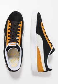 Svarta mockasneakers med gula och vita accenter, med en texturerad gummisula, vita sulor och svarta snören. PUMA-logotypen syns.