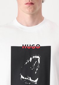 Hvit bomull T-skjorte med et grafisk trykk av en brølende munn i svart, med ordet "HUGO" i fete røde bokstaver over designet.
