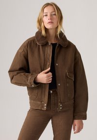 Vrouw in een bruine jas met schapenvachtkraag en bijpassende broek, die de voorkant van de jas met één hand vasthoudt, staand voor een effen lichte achtergrond.