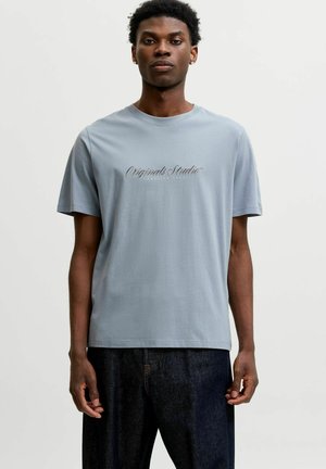 Jeune homme aux cheveux courts et bouclés portant un t-shirt bleu clair avec le texte "Originals Studio" et un jean foncé sur un fond uni.