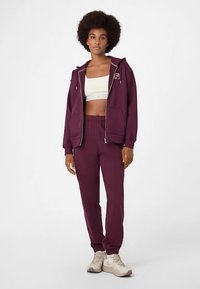 Bordeauxroter Kapuzenpullover mit Reißverschluss und Fronttaschen, passende Jogginghose und ein leichtes Tanktop. Verfügt über kontrastierende Nähte und elastische Bündchen.