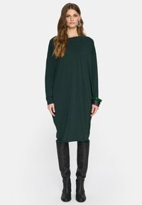 Kobieta ubrana w ciemnozieloną, luźną sukienkę z długim rękawem, czarne kozaki sięgające do kolan, duże kolczyki oraz kilka bransoletek na lewym nadgarstku.