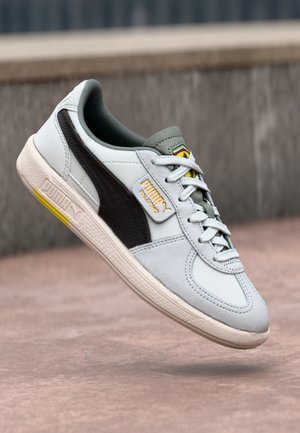 Weiß-grauer Puma-Sneaker mit schwarzem Streifen und gelben Akzenten, in der Luft über einer Betonfläche gezeigt.