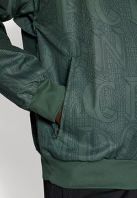 Green sweatshirt featuring an embossed logo pattern, zippered side pocket, ribbed cuffs, and a soft, textured fabric finish.