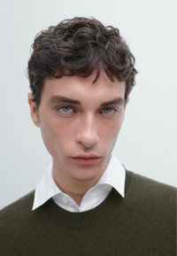 Zelený pletený svetr přes bílou košili s límečkem, strukturovaná tkanina, krátké kudrnaté vlasy a výrazné modré oči na neutrálním pozadí.