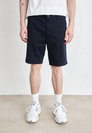 Man draagt een zwarte korte broek, witte sokken en witte Lacoste sneakers, staand op een vlakke vloer tegen een witte muur.