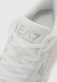 Fehér tornacipő texturált, párnázott talpbetéttel, fűzős kialakítással és az EA7 logó dombornyomott motívumával a nyelven és az oldalon, geometriai mintákkal díszítve.
