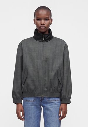 Person med barberet hoved iført en mørkegrå lynlåstrøje med sort krave og blå jeans, der står mod en ensfarvet lys baggrund.