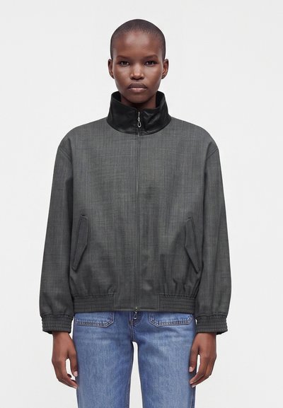 Personne à la tête rasée portant une veste zippée gris foncé avec un col noir et un jean bleu, debout devant un fond clair uni.