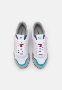 Sneakers bianche con materiali in mesh e suede, dettagli blu e rosa, suole texturizzate e lacci bianchi con dettagli di branding rossi e blu.