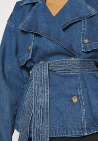 Jeansjacka i medelblått med bälte i midjan, bred krage och dubbelknäppning. Synlig sömnad och texturerad tygdetalj.