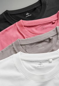 Vier Rundhals-T-Shirts gestapelt: schwarz, pink, grau und weiß. Jedes hat ein Etikett mit "DIE SET" und Pflegeanweisungen auf der Rückseite.