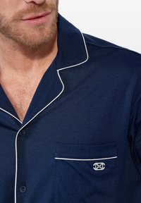 Camisa de pijama de manga corta en color azul marino, confeccionada en una tela suave, con ribete blanco en el cuello y en el bolsillo, además de un logo en el bolsillo del pecho.