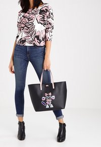 Kvinna klädd i blommig rosa och vit topp och blå jeans, som håller en svart tote-väska med färgglad geometrisk kattdesign och rosa handtag.