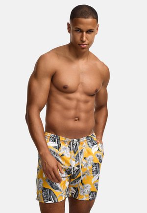 Homme musclé aux cheveux courts portant un short de bain jaune avec des motifs de feuilles et de fleurs tropicales blanches et noires, mains dans les poches.