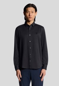 Camicia nera a maniche lunghe con bottoni, caratterizzata da un motivo testurizzato, bottoni azzurri e un piccolo logo sul petto. Indossata con pantaloni scuri.