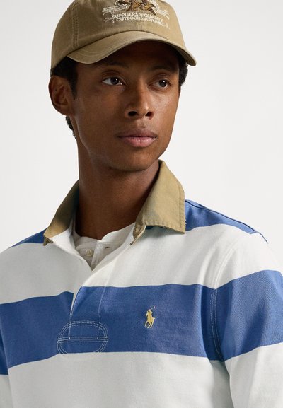 Muž v baretke béžovej farby a bielom ragby tričku s modrými horizontálnymi pruhmi a béžovým golierom, hľadí mierne do strany.