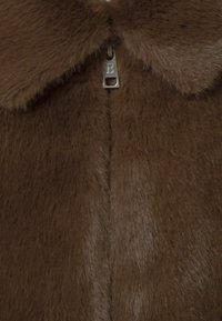 Abrigo marrón de piel sintética con textura suave, que cuenta con un cuello redondeado y un cierre de cremallera adornado con una etiqueta metálica marcada con una "B".