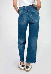 Jeans de denim azul con un corte relajado, con bolsillos traseros y una etiqueta de cuero en la cintura, combinados con zapatillas blancas.