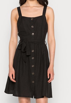 Femme portant une robe noire sans manches à boutons sur le devant, avec un col carré et une ceinture nouée à la taille, debout devant un fond uni.