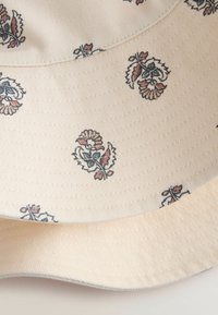 Een bucket hat van beige stof met een gestructureerde afwerking, versierd met bloemmotieven in blauw en rood. De stiksels en de rand zijn duidelijk zichtbaar.