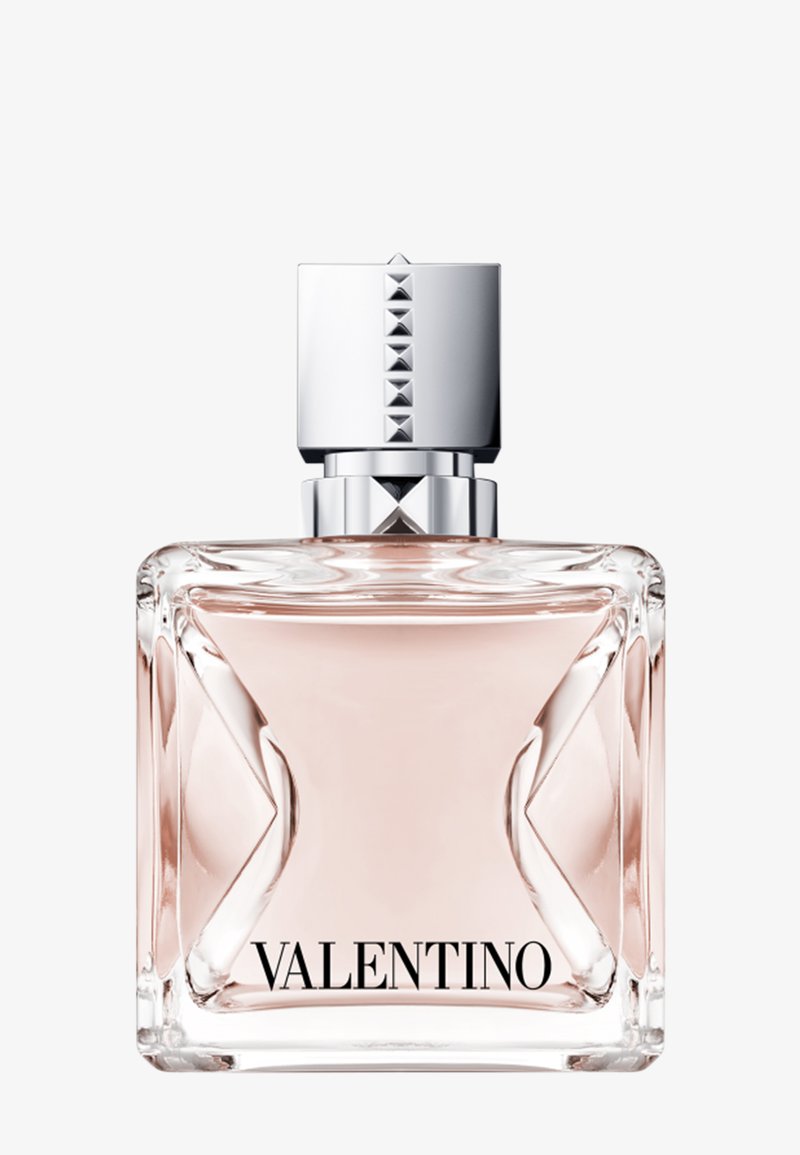 Flacon de parfum en verre transparent avec un liquide rose pâle, bouchon argenté orné de clous pyramidaux, et "VALENTINO" imprimé en bas.